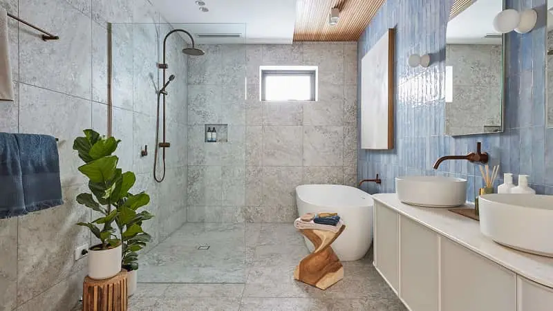 Modern bathroom with marble tiles, walk-in shower, freestanding tub, double sinks, and a green plant on wooden stand.