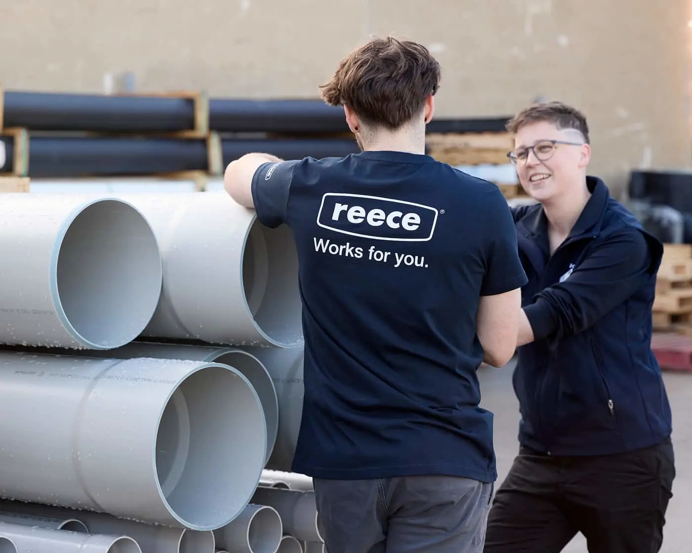 Two workers in navy uniforms near large PVC pipes, one wearing a Reece shirt with "Works for you" slogan.