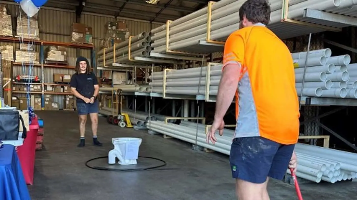 Two workers in a warehouse with PVC pipes stacked on shelves, playing a game with a toilet in the center.