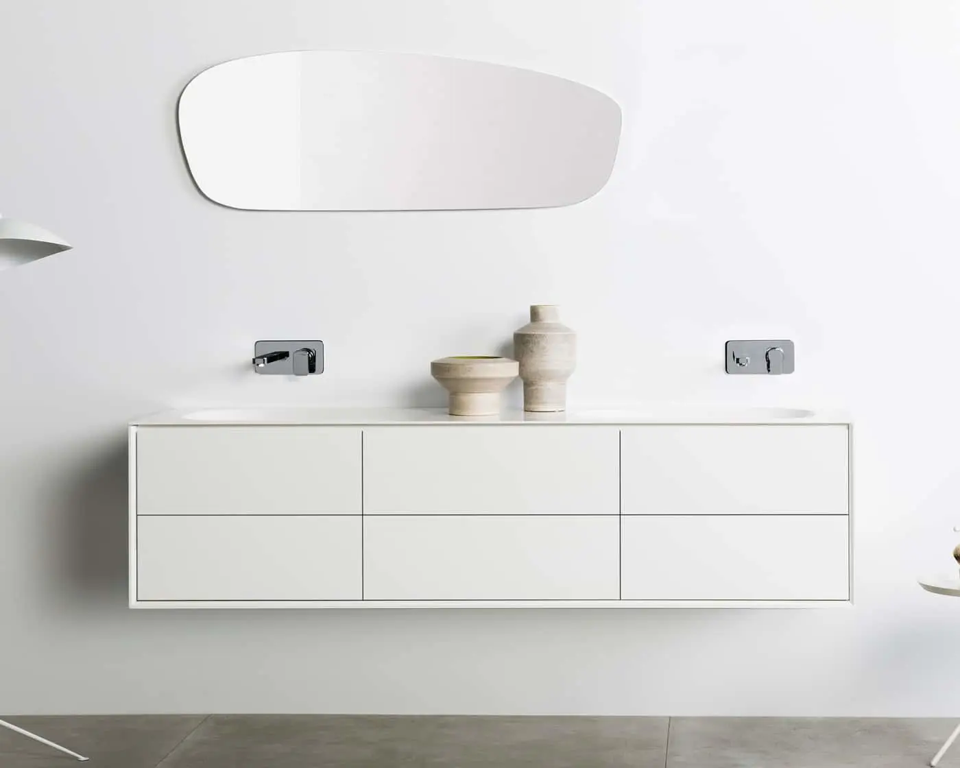 Minimalist white bathroom vanity with wall-mounted faucets, ceramic decorative vessels, and oval mirror above.