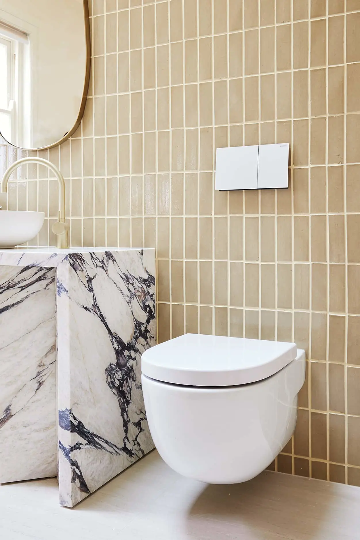 Modern bathroom with beige tile walls, white wall-mounted toilet, marble vanity, round mirror, and flush button.
