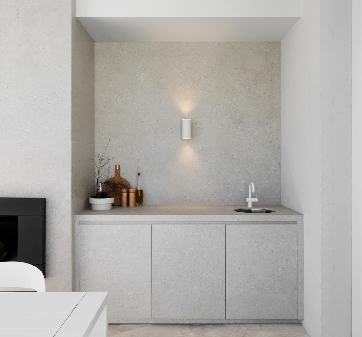 Minimalist kitchen alcove with concrete countertop, white wall sconce, sink, and decorative wooden containers.