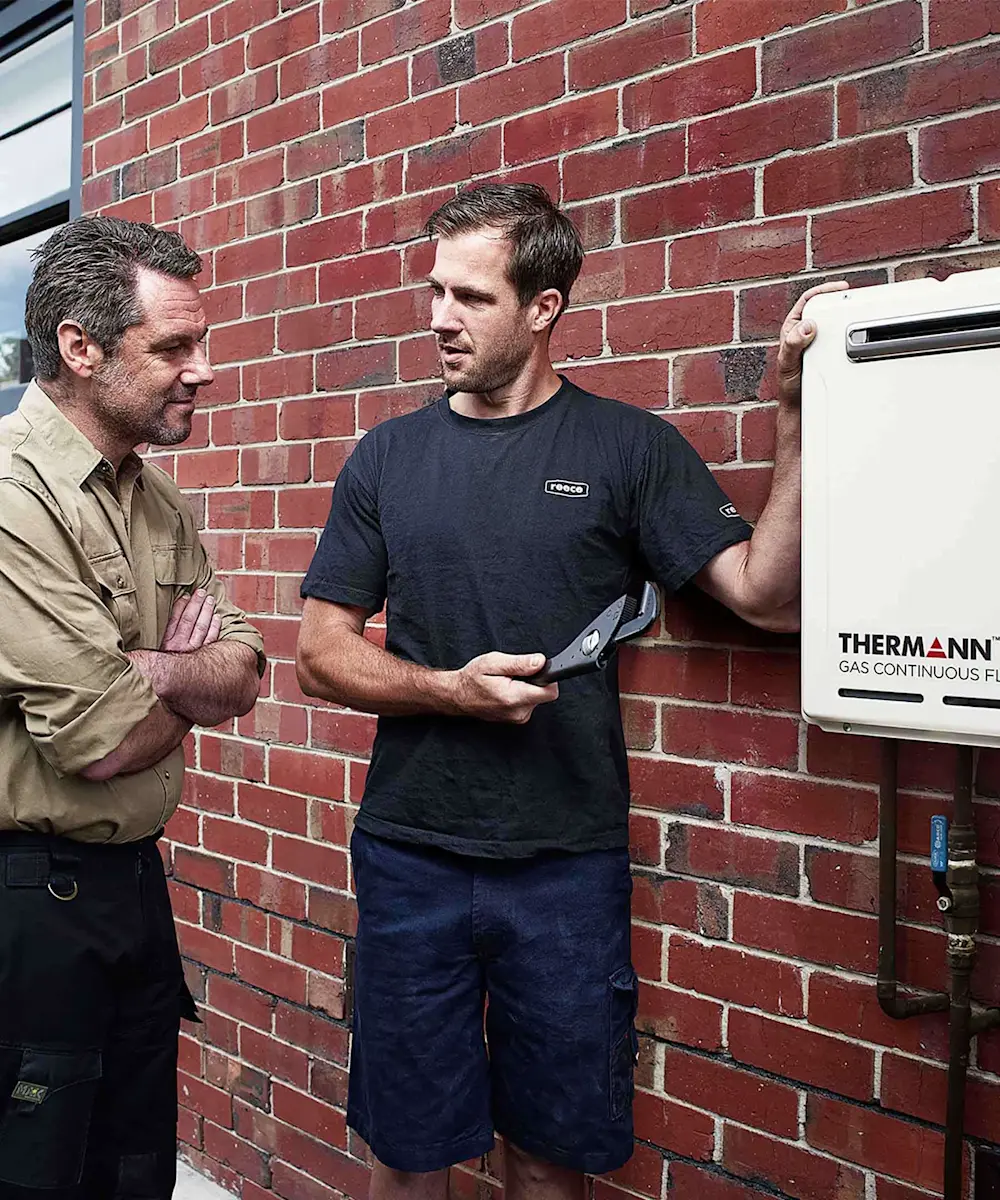 Two men discussing next to a Thermann gas system mounted on a red brick wall, one holding a tool.