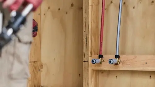 Red and blue plumbing pipes with connectors mounted on wooden framing inside an unfinished wall.