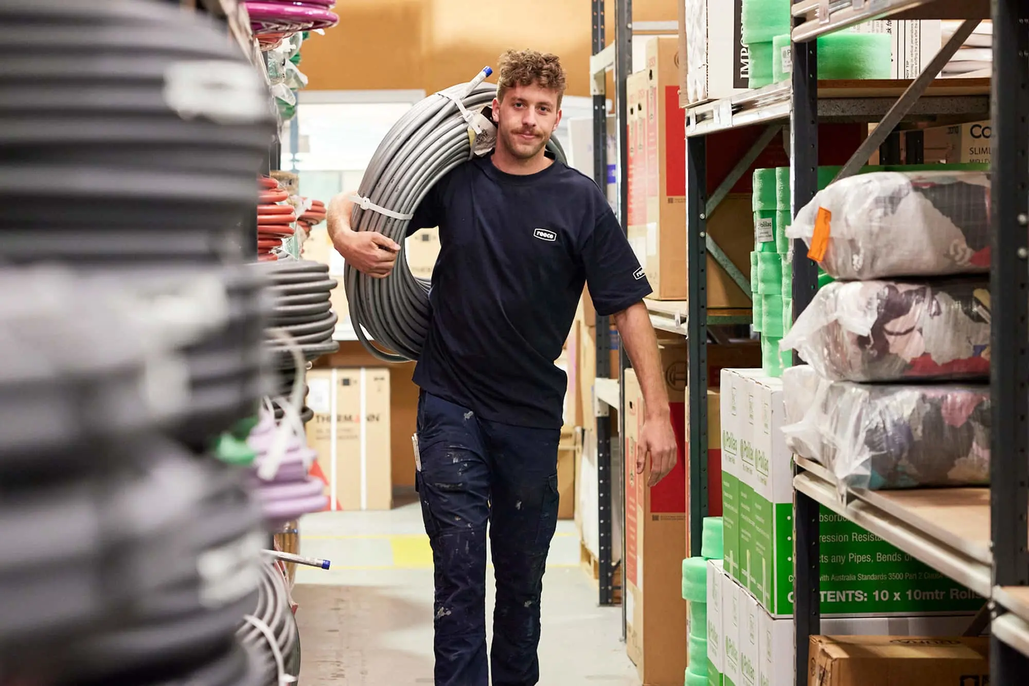 Worker in navy uniform carrying coiled tubing while walking through warehouse shelves stocked with construction supplies.