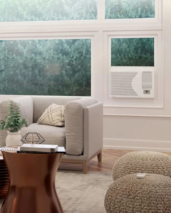 Modern living room with beige armchair, window air conditioner, wooden side table, and textured pouf on hardwood floor.