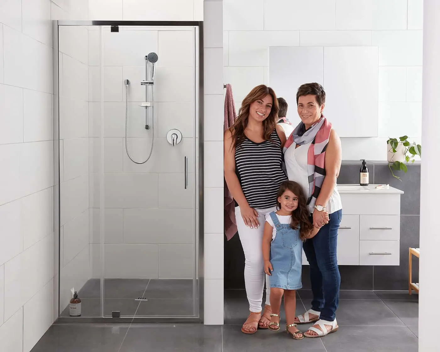 Modern bathroom with glass shower enclosure and three people standing together beside white vanity cabinet.