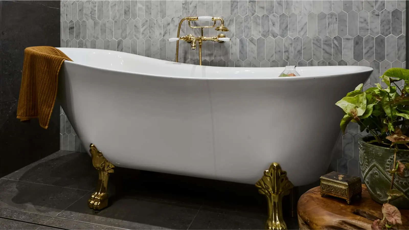 White clawfoot bathtub with gold fixtures against gray hexagonal tile wall, orange towel and potted plant nearby.