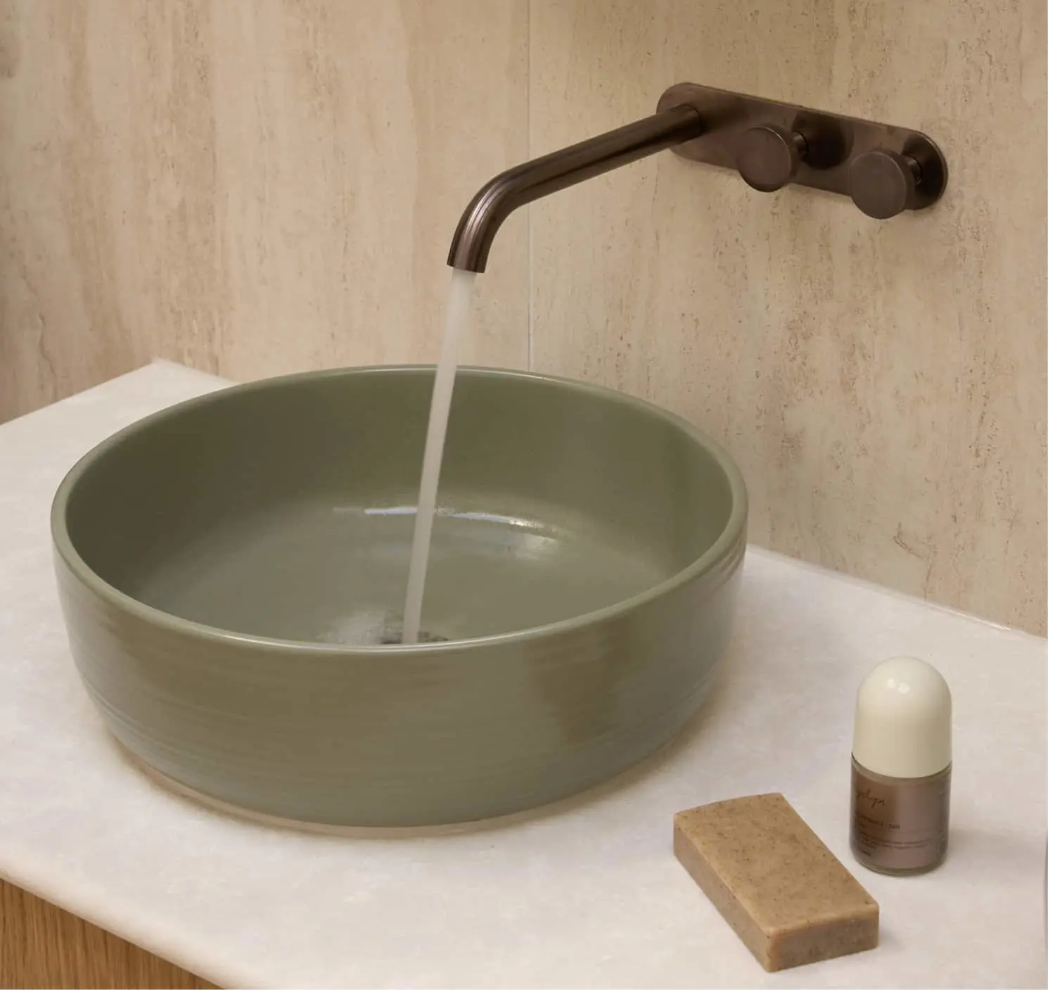 Wall-mounted bronze faucet filling a round green ceramic vessel sink on a white countertop with soap and bottle.