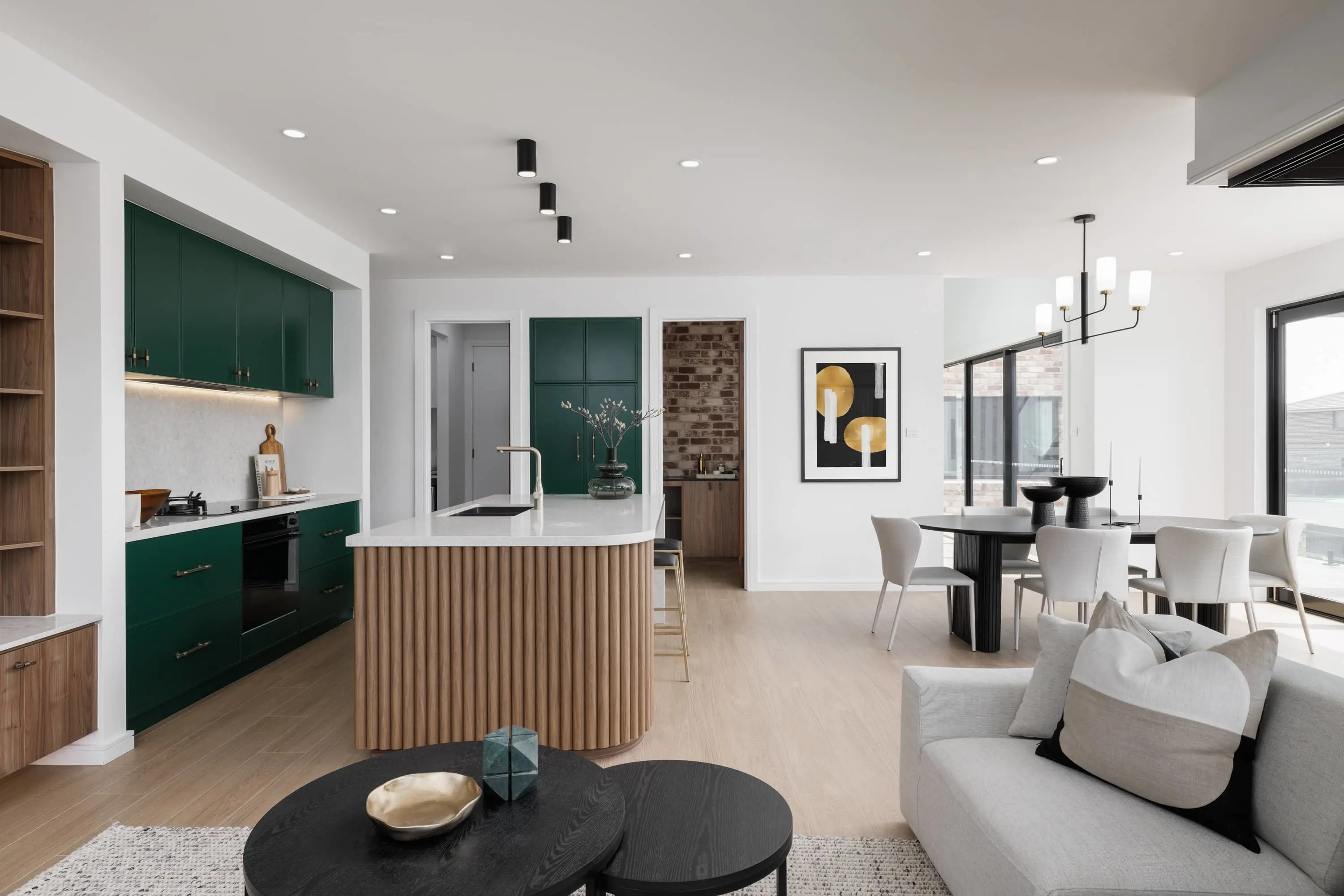 Modern open-concept kitchen and living area with dark green cabinets, wooden island, white dining table, and light gray sofa.