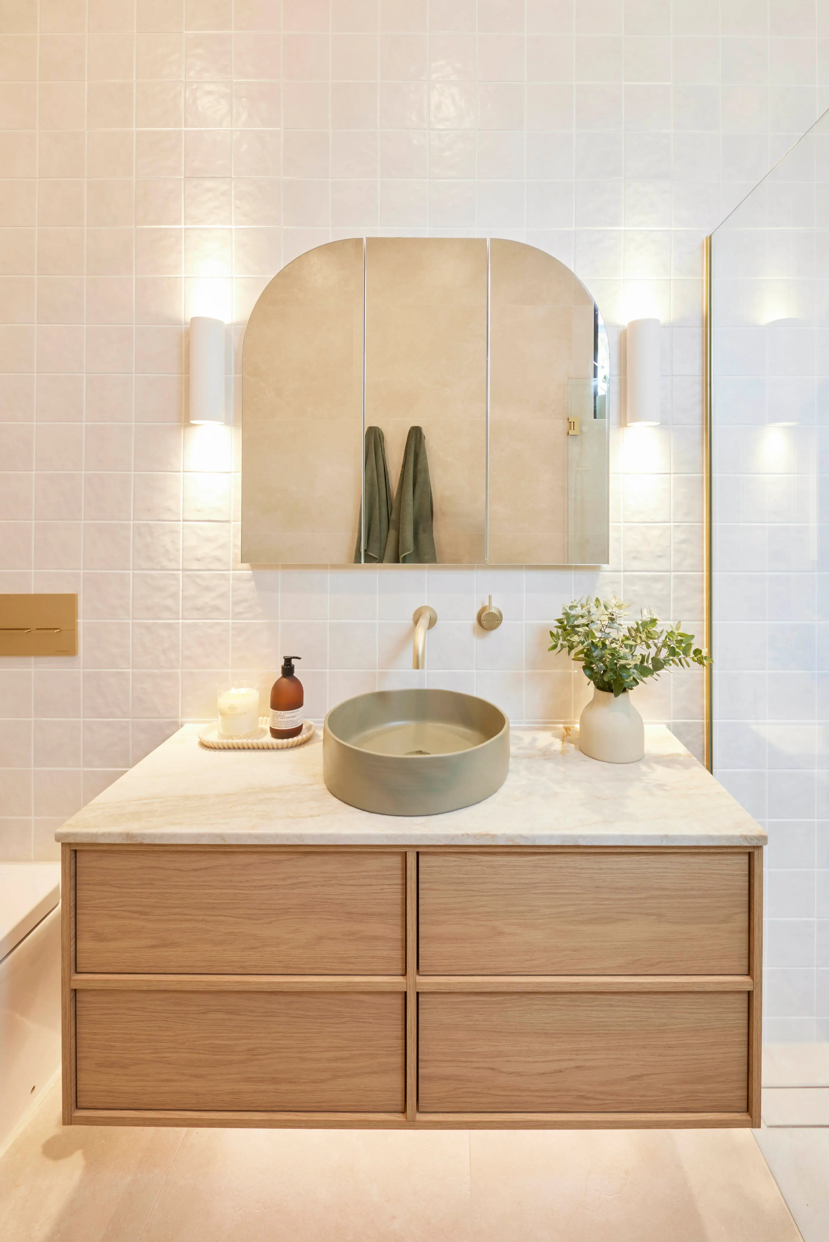 Modern bathroom with wooden vanity, round sink, arched mirror, white tiled walls, and small plant in vase.