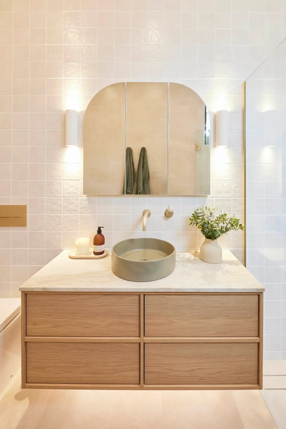 Modern bathroom with wooden vanity, round sink, arched mirror, white tiled walls, and small plant in vase.