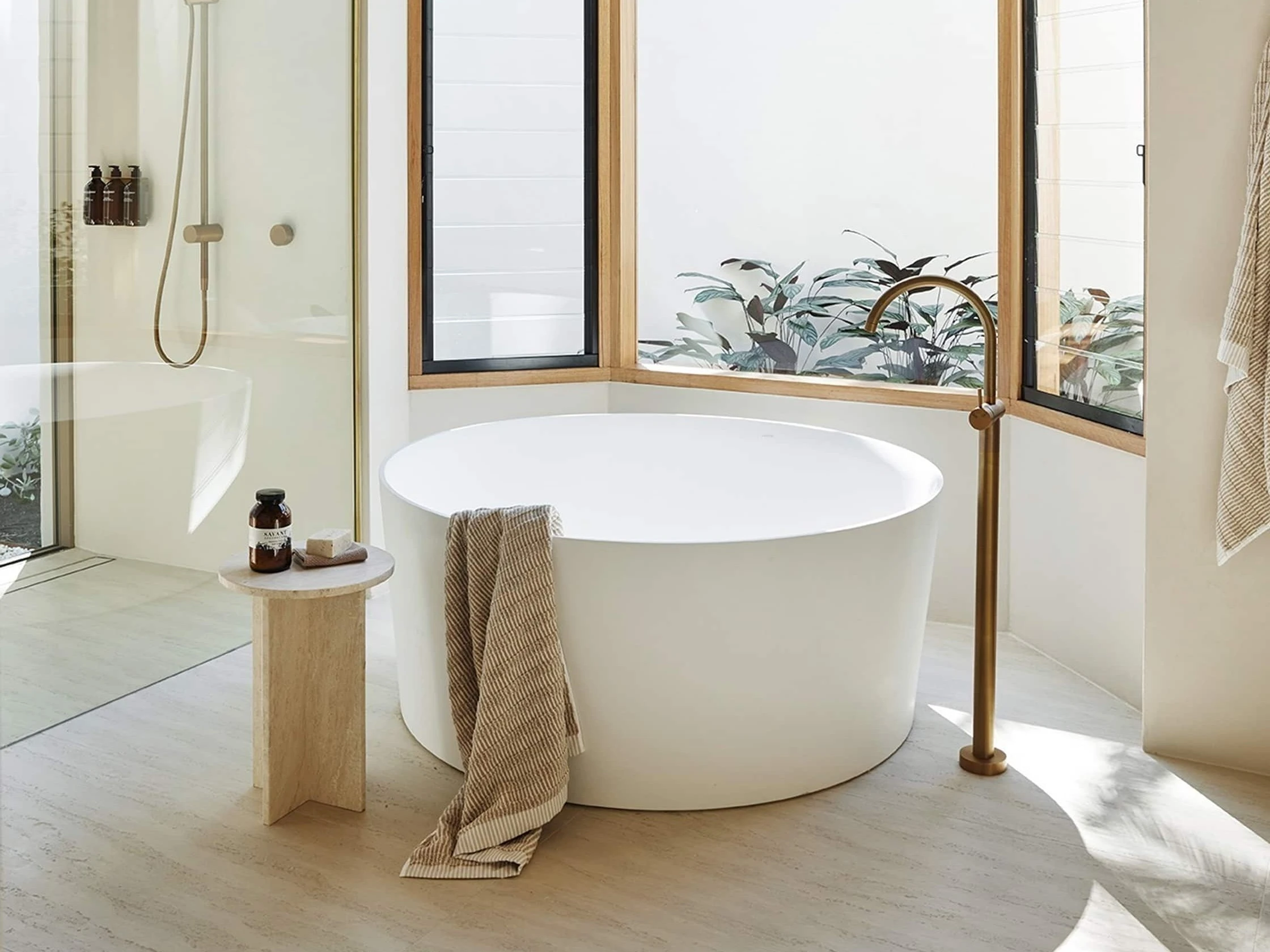 Modern bathroom with white round tub, glass shower, and large windows overlooking tropical plants and palm trees.