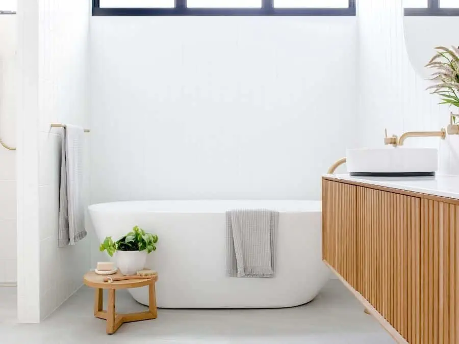 Modern white bathroom with freestanding tub, wooden vanity, small plant on stool, and gray towels under windows.