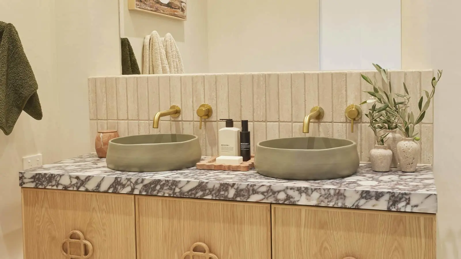 Modern bathroom with dual green vessel sinks, marble countertop, brass fixtures, and decorative plants on wooden vanity.