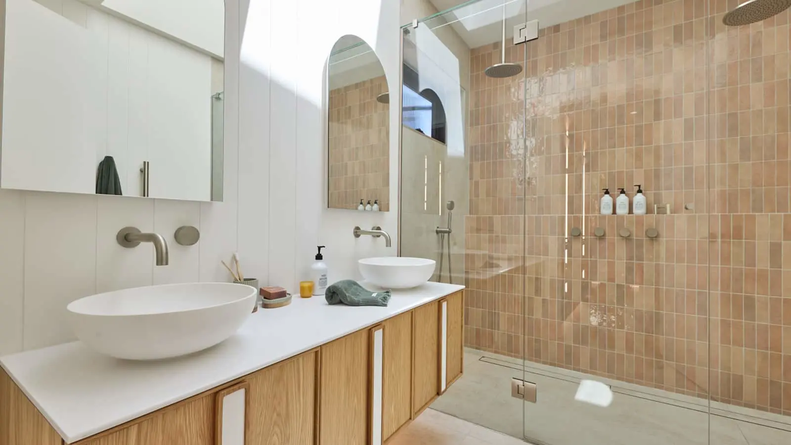 Modern bathroom with white vessel sinks, wooden vanity, and glass shower with peach-colored tile walls.