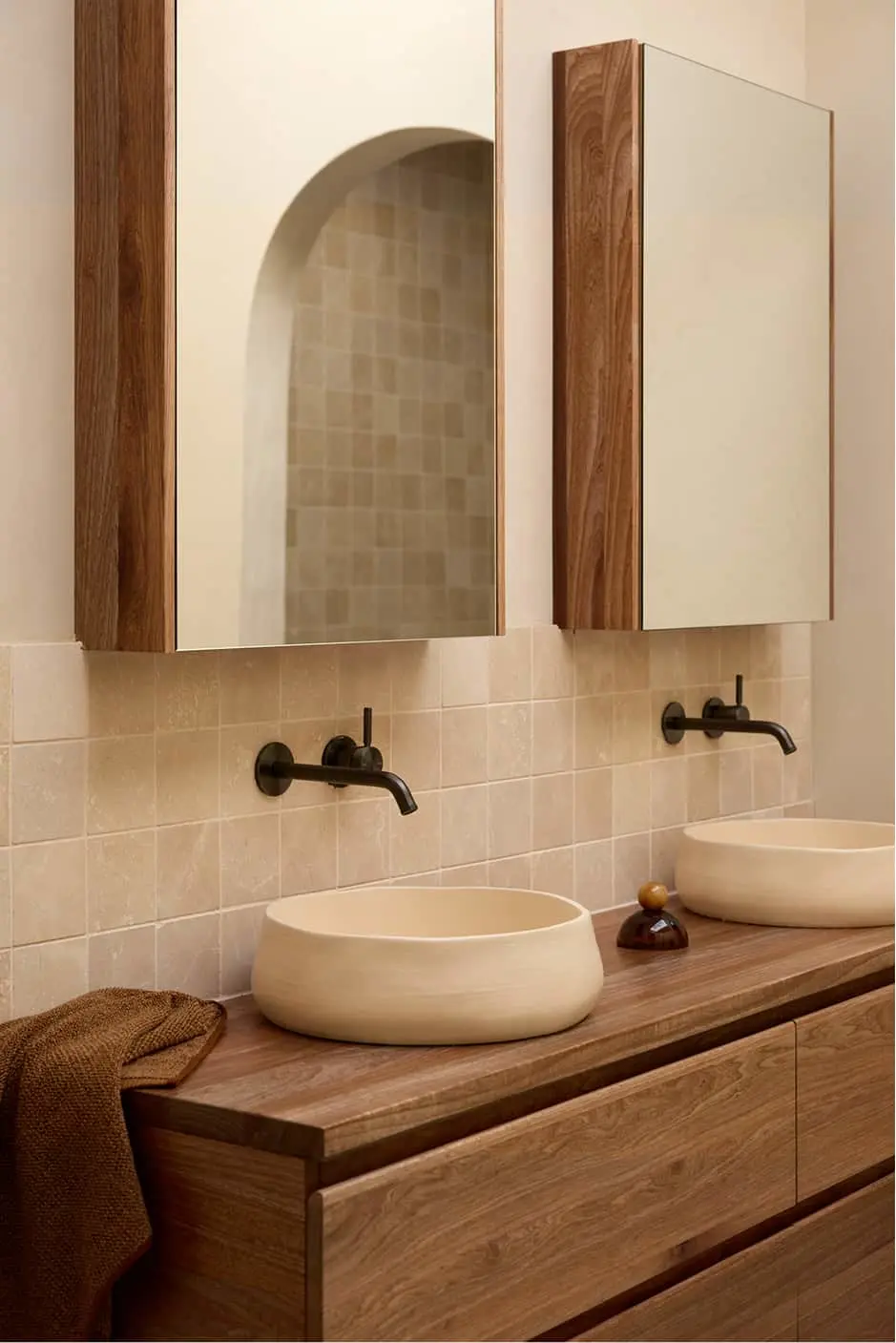 Modern bathroom with two cream vessel sinks on wood vanity, beige tile backsplash, black wall faucets, and wood-framed mirrors.
