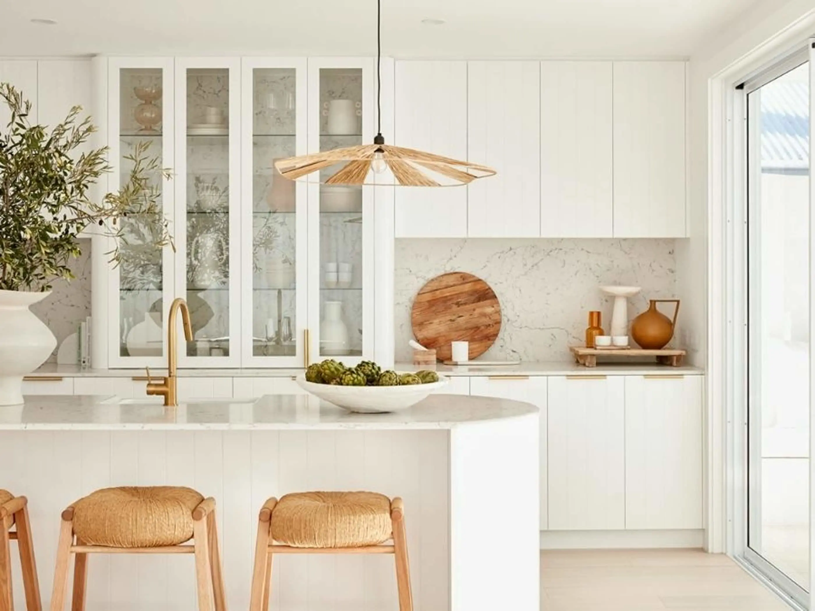 Modern white kitchen with marble countertops, woven pendant light, glass cabinets, and natural wood accents.