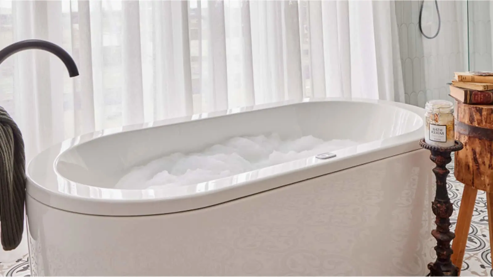 White freestanding bathtub filled with bubbles near sheer curtains, with wooden side table holding books and bath products.
