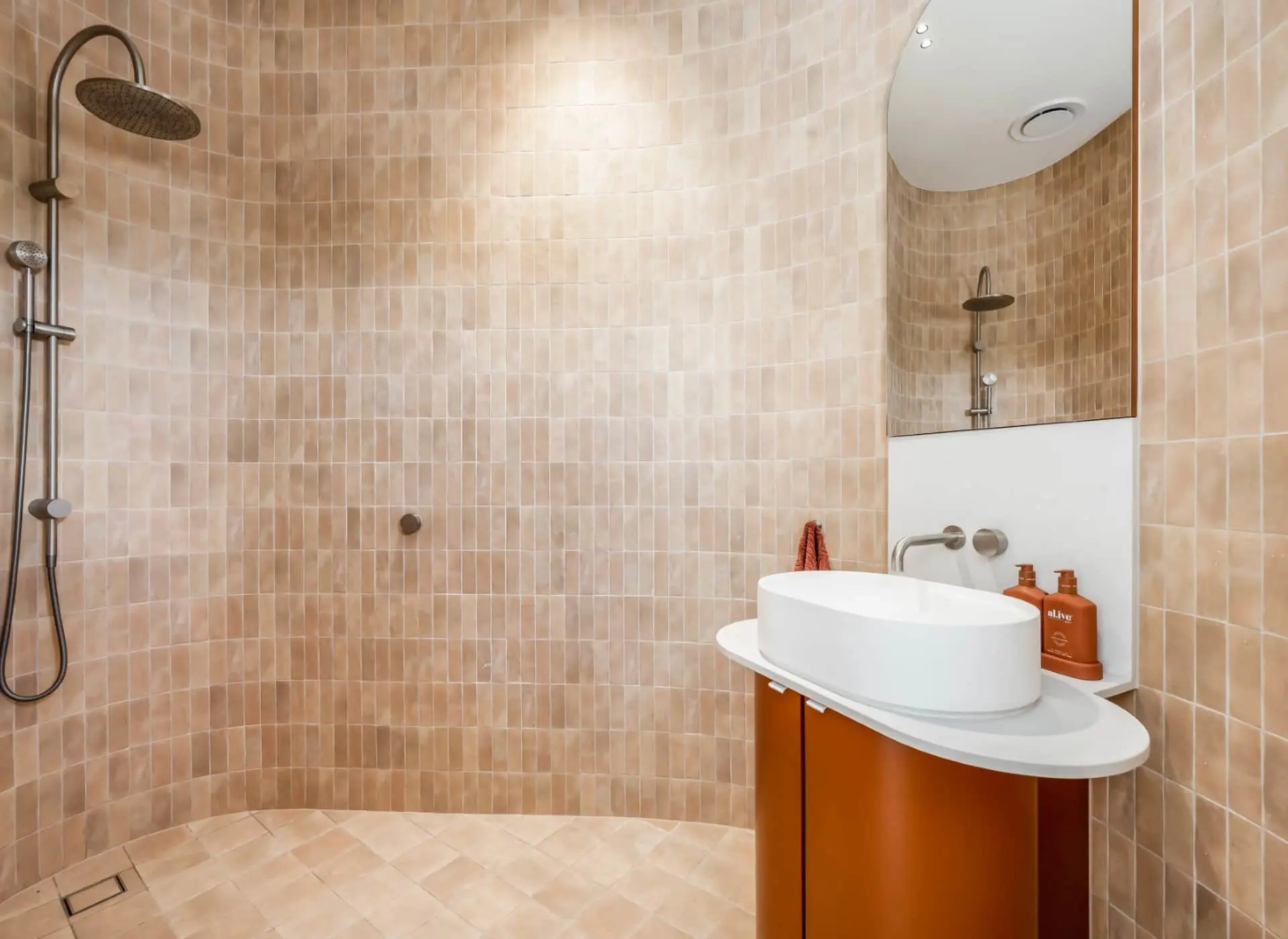 Modern bathroom with beige tile walls, rainfall shower, white oval sink on orange cabinet, and mirror.
