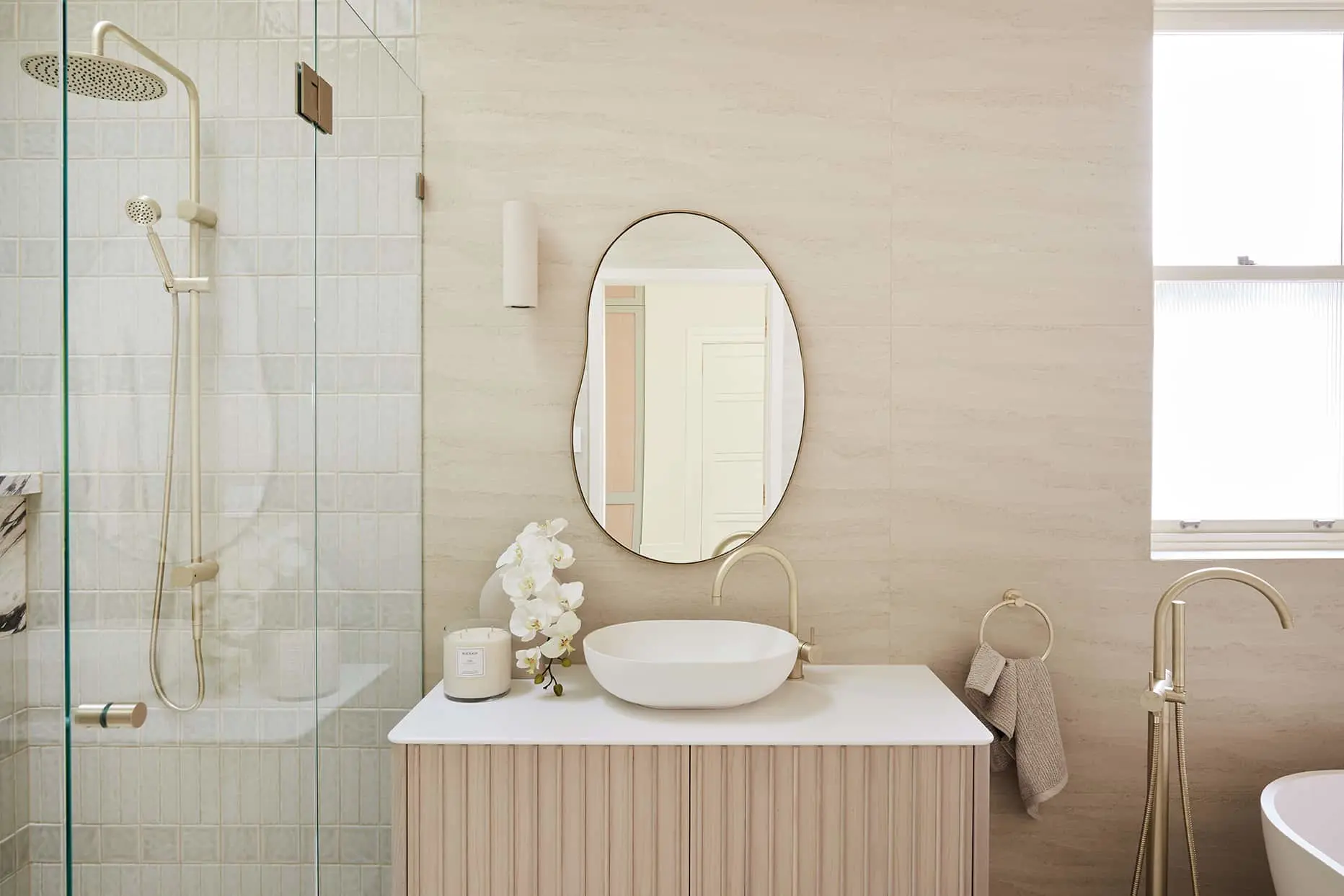 Modern bathroom with glass shower, oval mirror, white vessel sink, and beige tones featuring gold fixtures and white orchids.