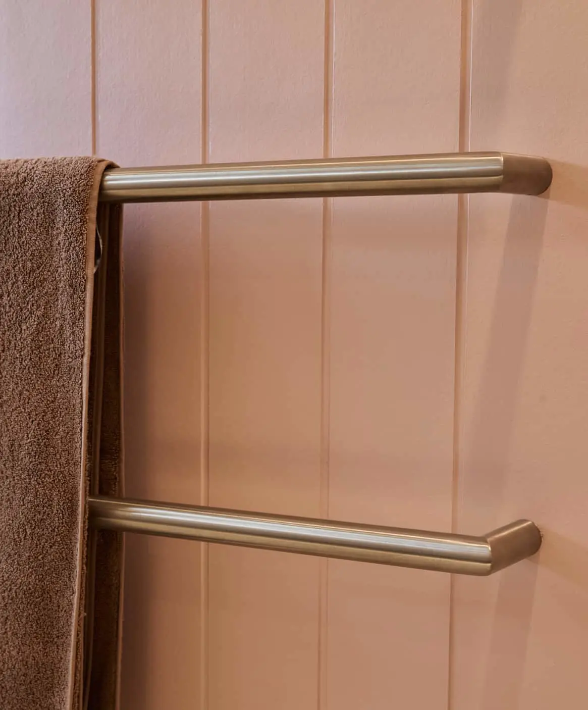 Gold double towel bar mounted on a light pink paneled wall with a brown towel hanging on the left side.