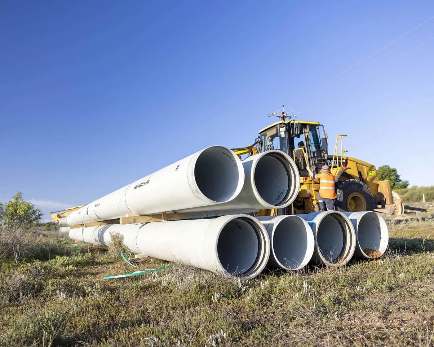 Yellow construction loader moving large white drainage pipes at a construction site under clear blue sky.