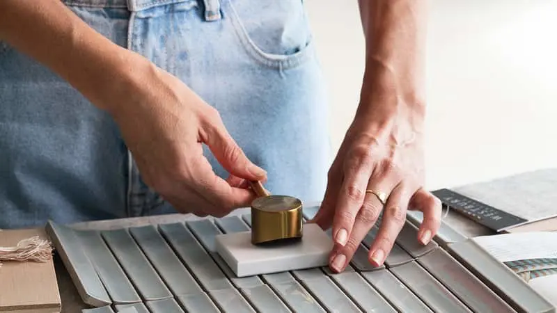 Person in light jeans placing a small gold cylinder on a white tile over metal samples on a workspace.