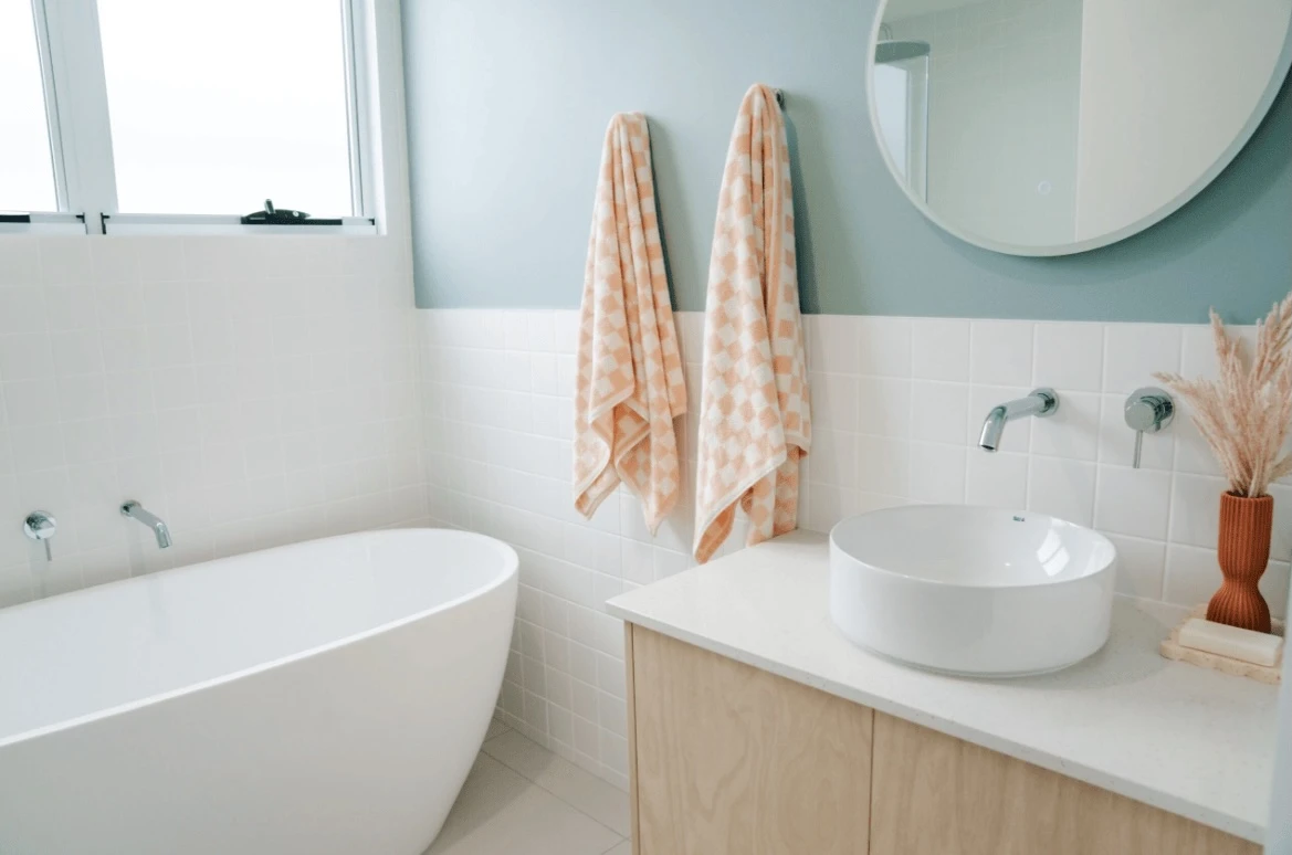 Modern bathroom with white bathtub, vessel sink, peach patterned towels, round mirror, and light wood vanity.