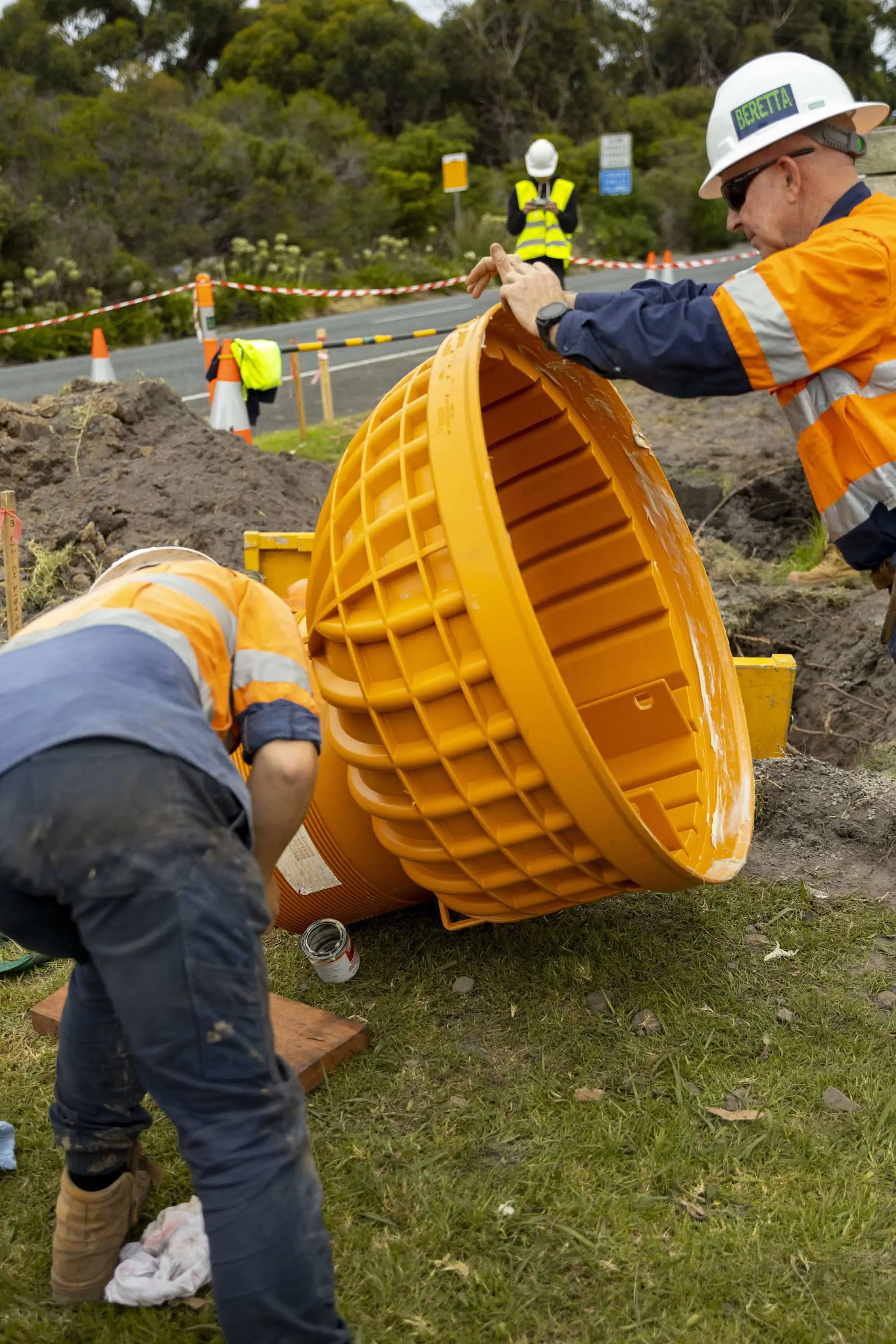 Construction workers installing a large yellow drainage pipe at a roadside worksite with safety barriers and traffic control.