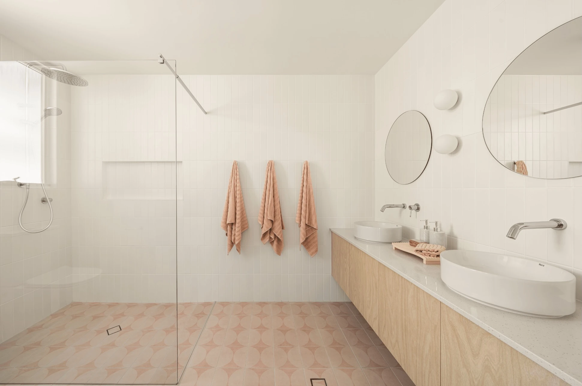 Modern white bathroom with glass shower, peach towels, wooden vanity with two sinks, and patterned pink floor tiles.