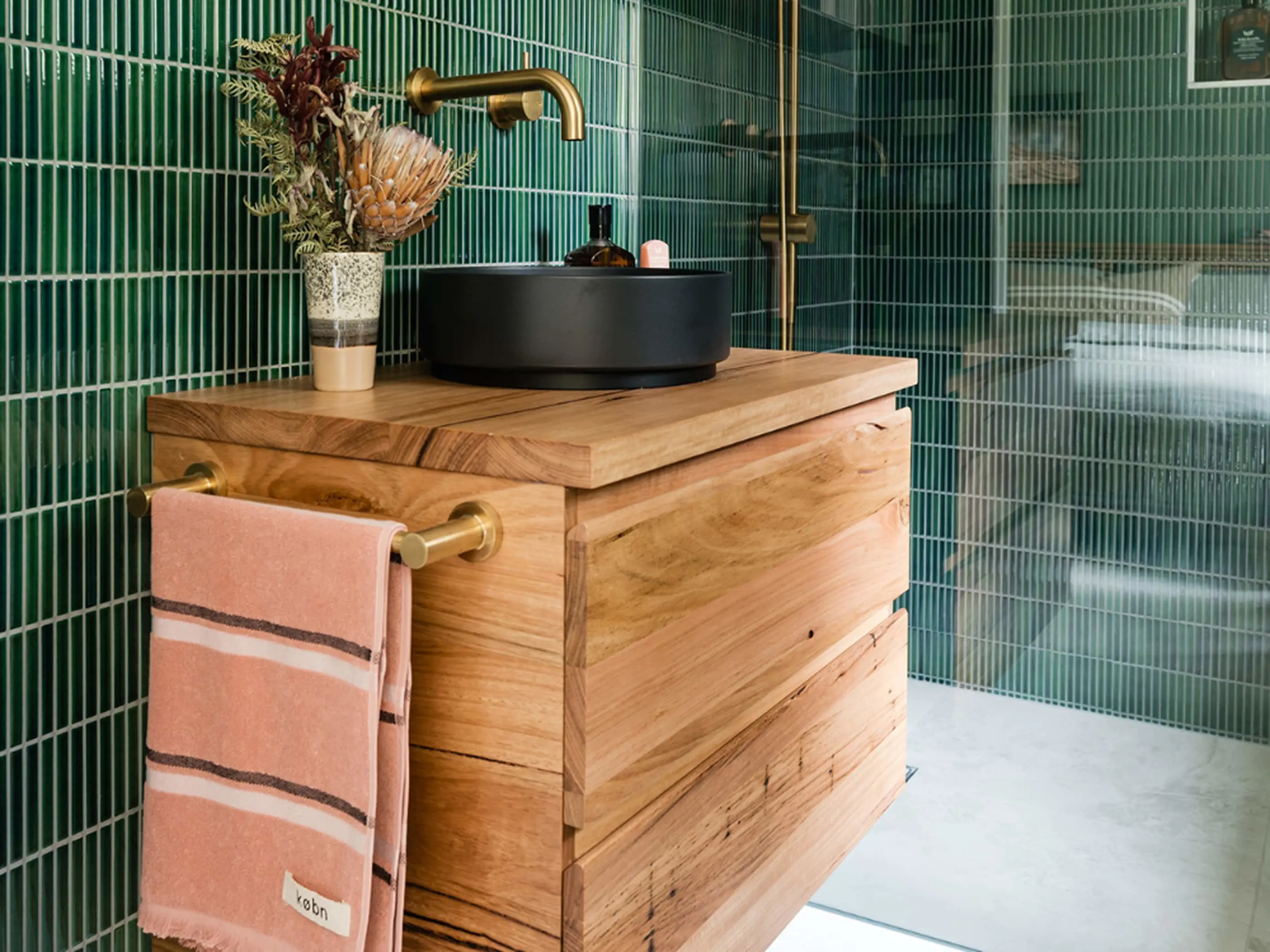 Modern bathroom with wooden vanity, black basin, brass fixtures, green tiled walls, and pink towel.