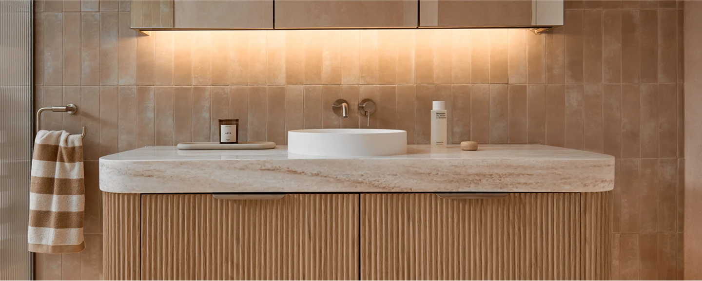 Modern bathroom with beige tiled walls, marble countertop, white vessel sink, and wooden cabinetry with striped towel hanging nearby.