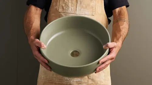 Person in tan apron holding a round sage green ceramic sink basin with smooth finish and center drain hole.