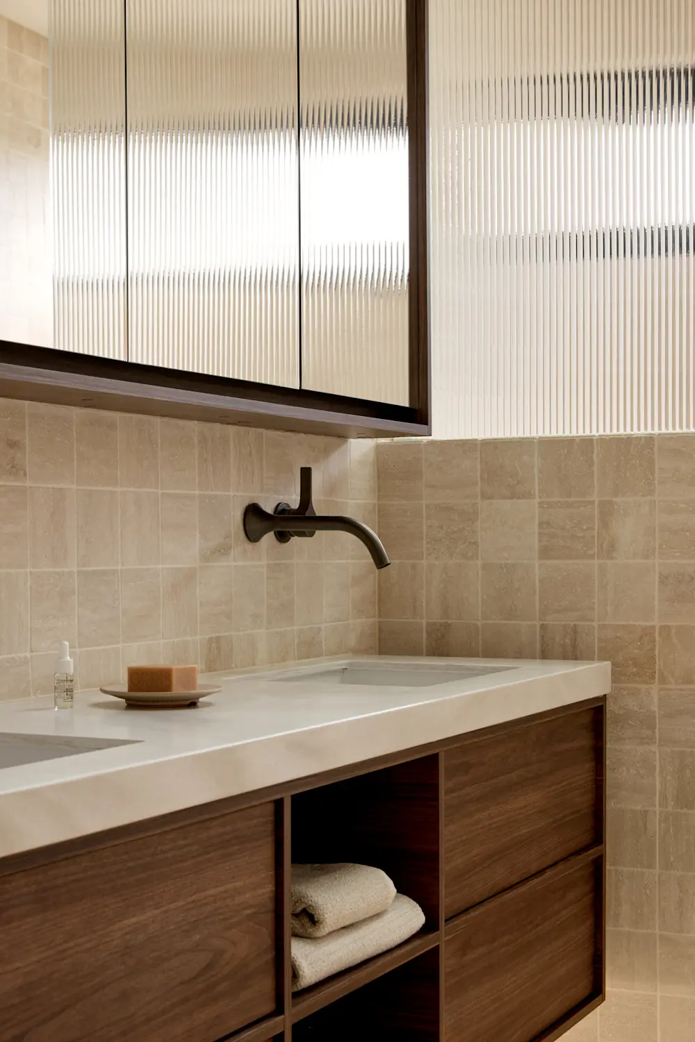 Modern bathroom with wooden vanity, white countertop, wall-mounted faucet, and ribbed glass cabinet above beige tiled walls.