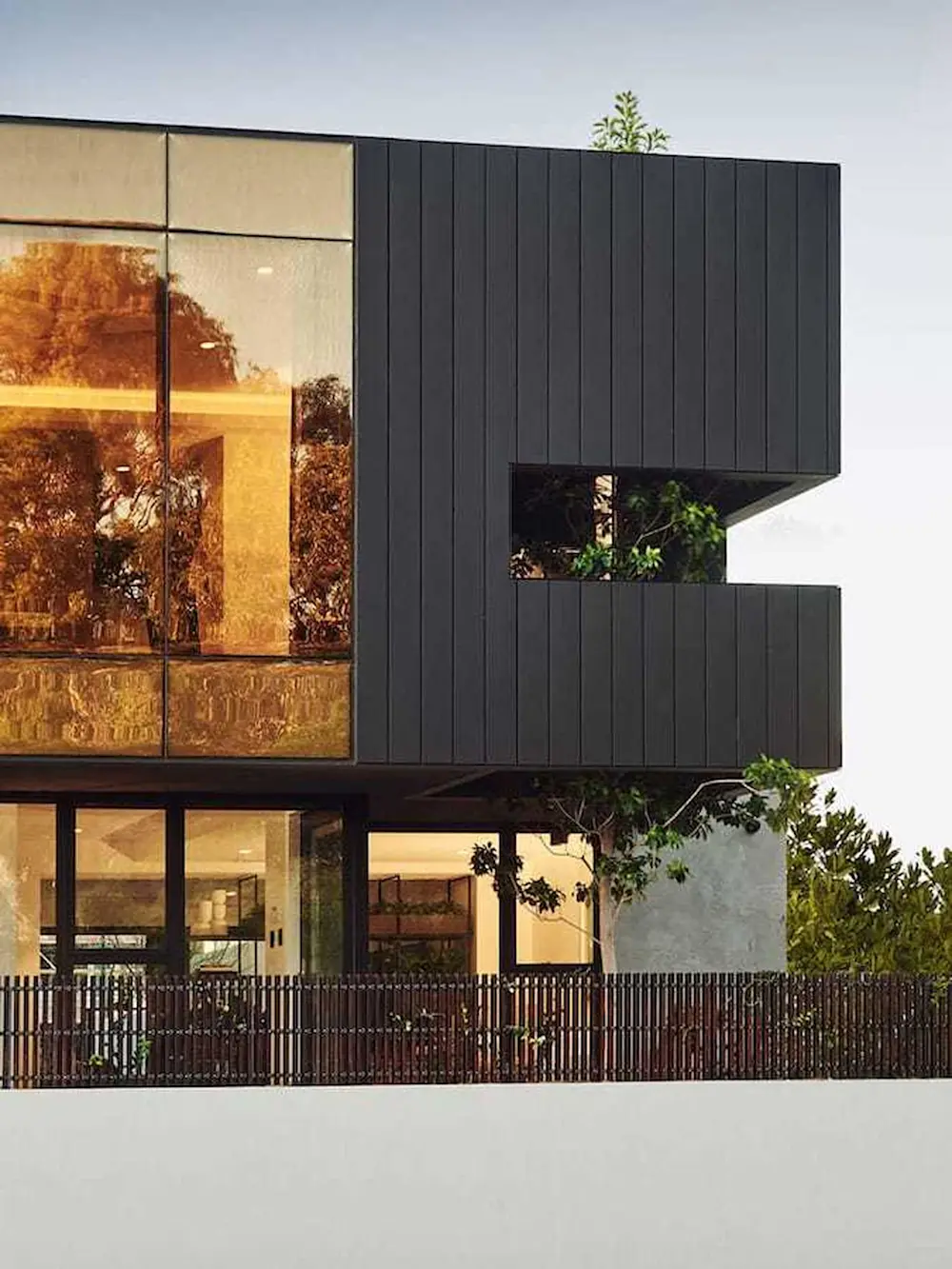 Modern house with black metal exterior, large glass windows reflecting warm light, and a metal fence surrounding the property.