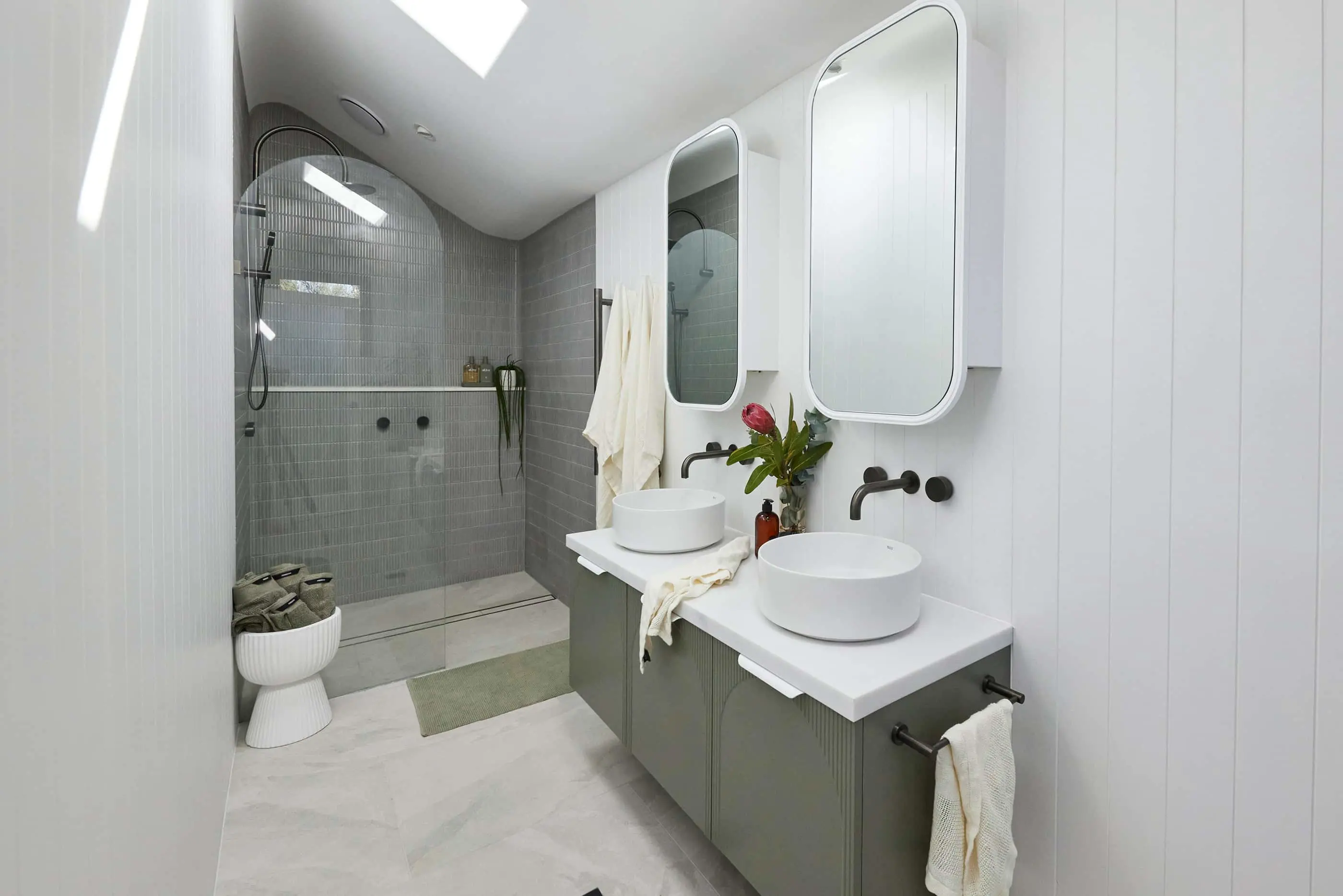 Modern bathroom with gray tiled shower, dual white vessel sinks, rounded mirrors, and a small vase with red flower.