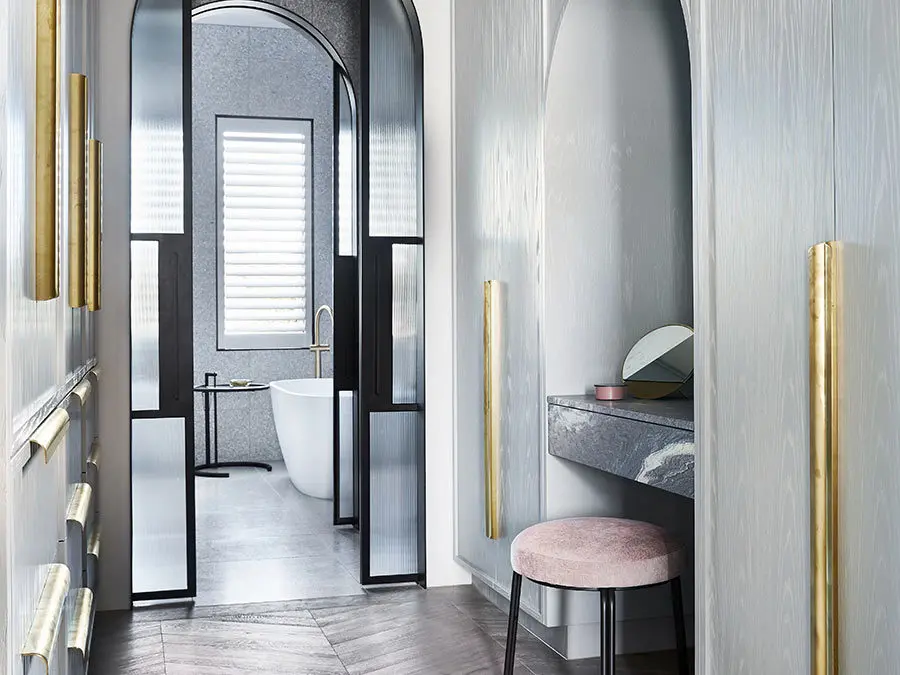Modern bathroom entrance with arched doorway, freestanding tub, pink stool, and gold accents on light gray walls.