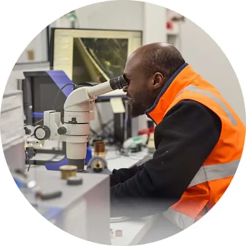 Worker in orange safety vest looking through microscope in laboratory setting.