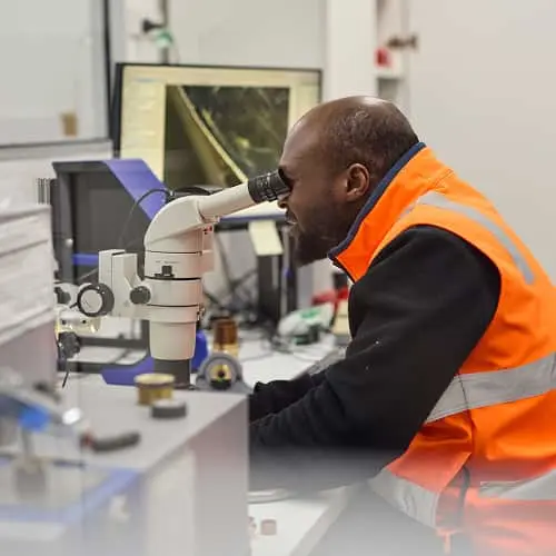 Worker in orange safety vest looking through microscope in laboratory setting.