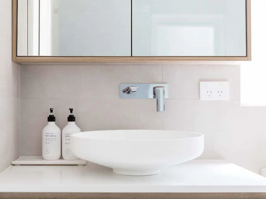 Modern bathroom sink with white vessel basin, wall-mounted faucet, wooden mirror cabinet, and two soap dispensers.