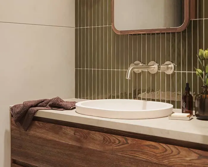 Modern bathroom with green vertical tiles, wooden vanity, white vessel sink, wall-mounted brass faucet, and small plant.