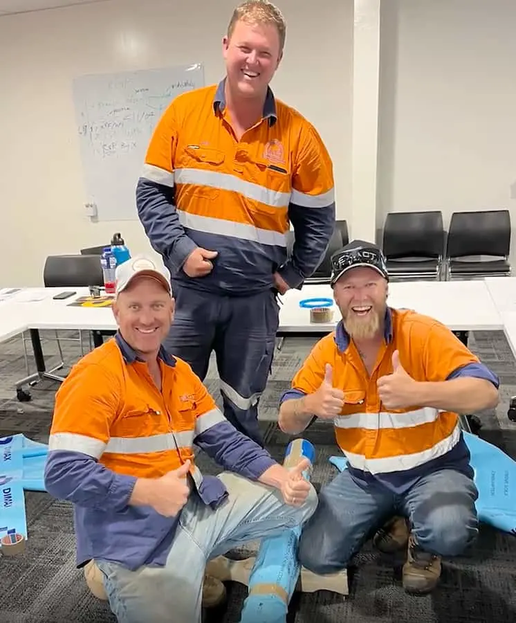 Three workers in orange and navy hi-vis uniforms smiling in a meeting room, two giving thumbs up.