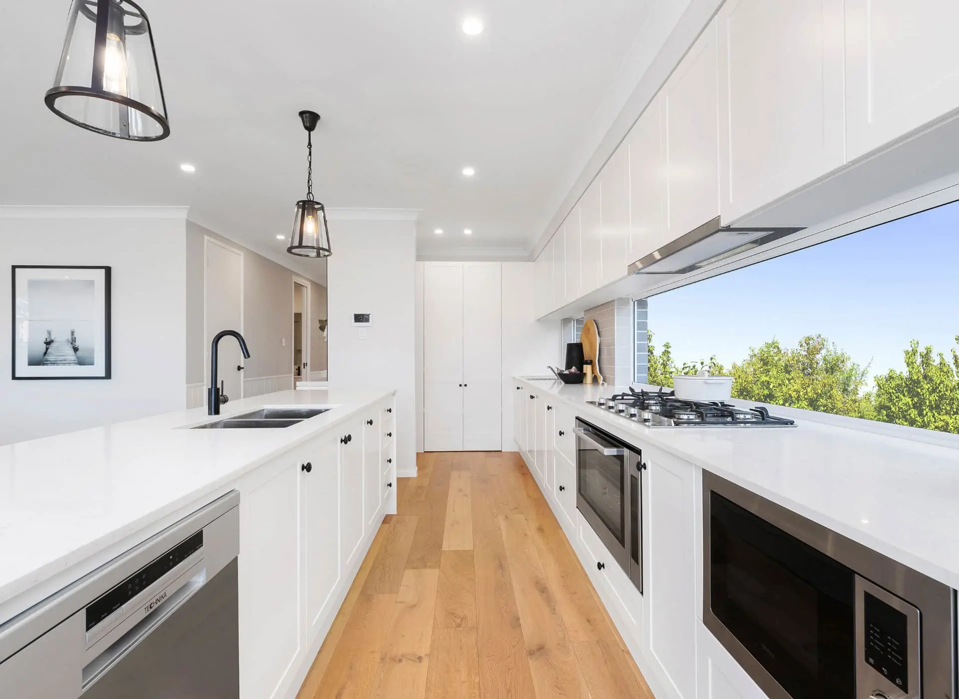 Modern white kitchen with wooden floor, pendant lights, large window overlooking greenery, and stainless steel appliances.