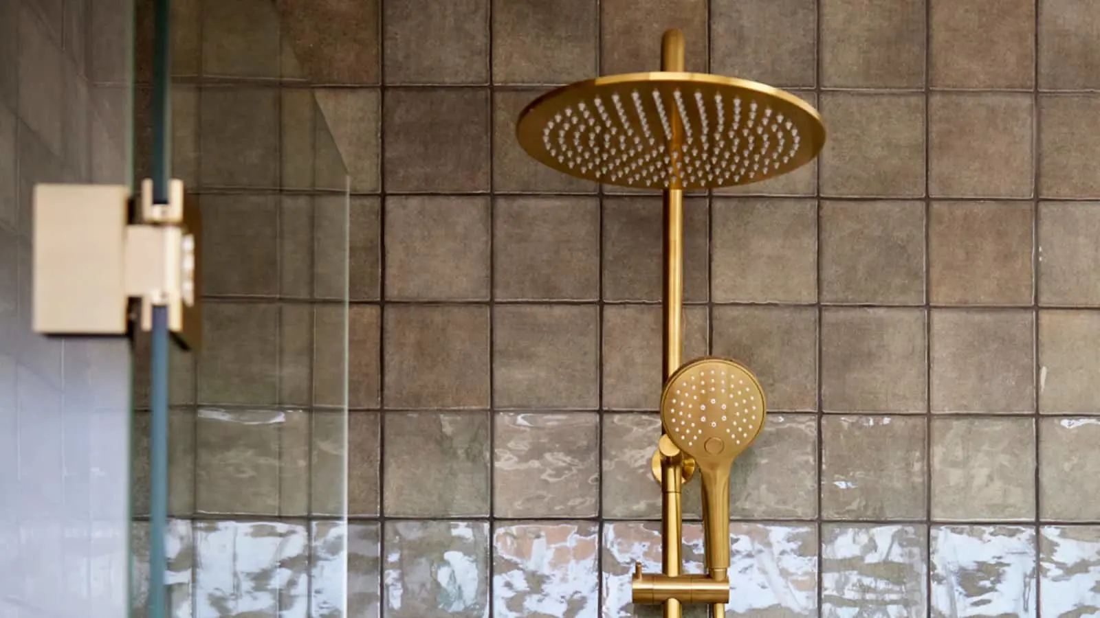Gold rainfall showerhead with handheld attachment mounted on gray tiled bathroom wall next to glass shower door.