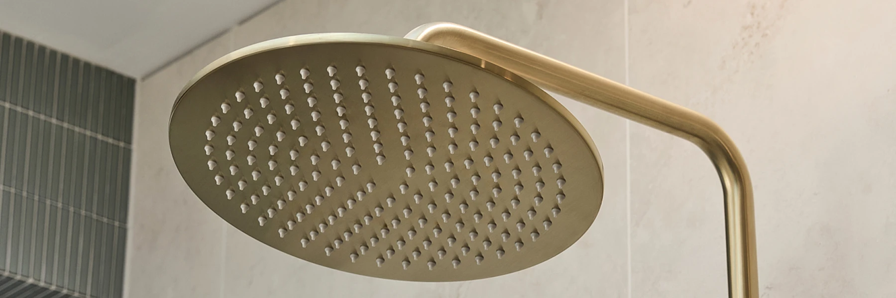 Round gold rainfall showerhead mounted on a curved arm against white bathroom tiles with green accent wall.