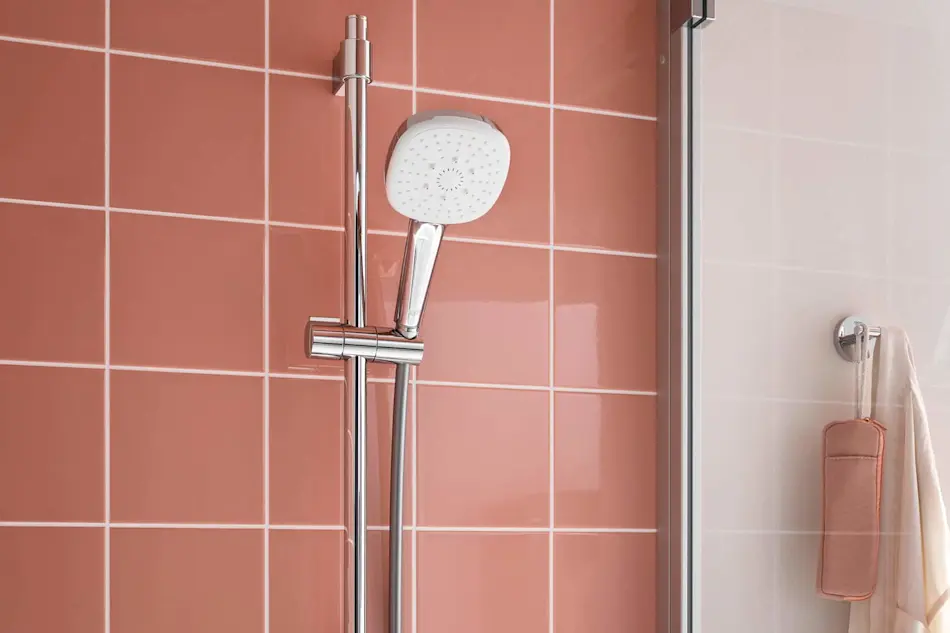 Modern shower head with adjustable rail mounted on coral pink tiled wall, with shower accessories hanging nearby.