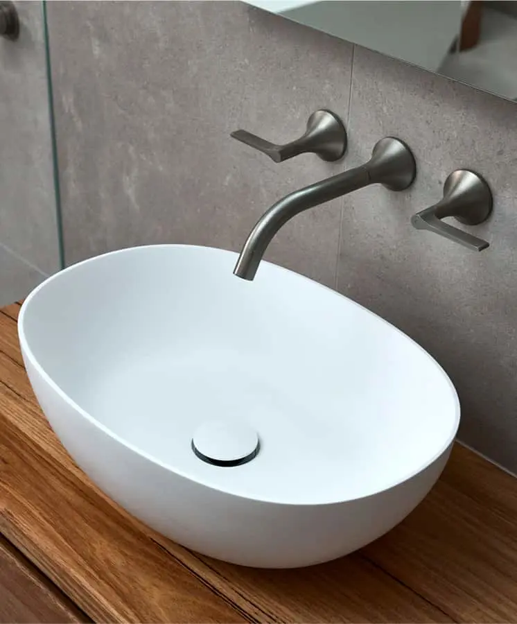 Modern white oval basin sink on wooden countertop with wall-mounted brushed metal faucet against gray tile wall.
