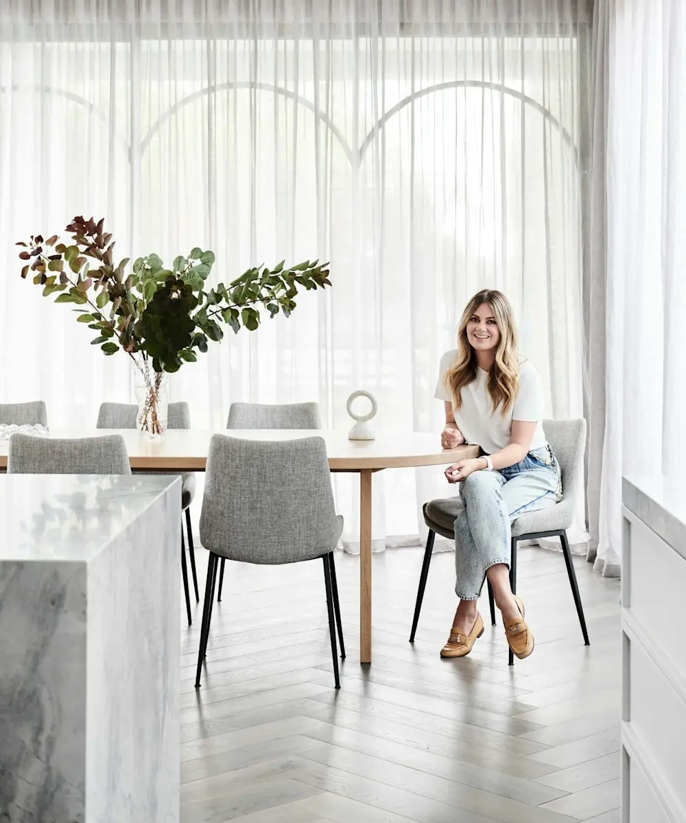Woman sitting at wooden dining table with gray chairs, greenery centerpiece, and sheer white curtains in bright room.
