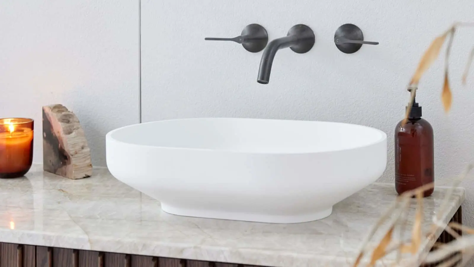 Modern bathroom with white vessel sink, black wall-mounted faucet, marble countertop, lit candle and decorative bottle.